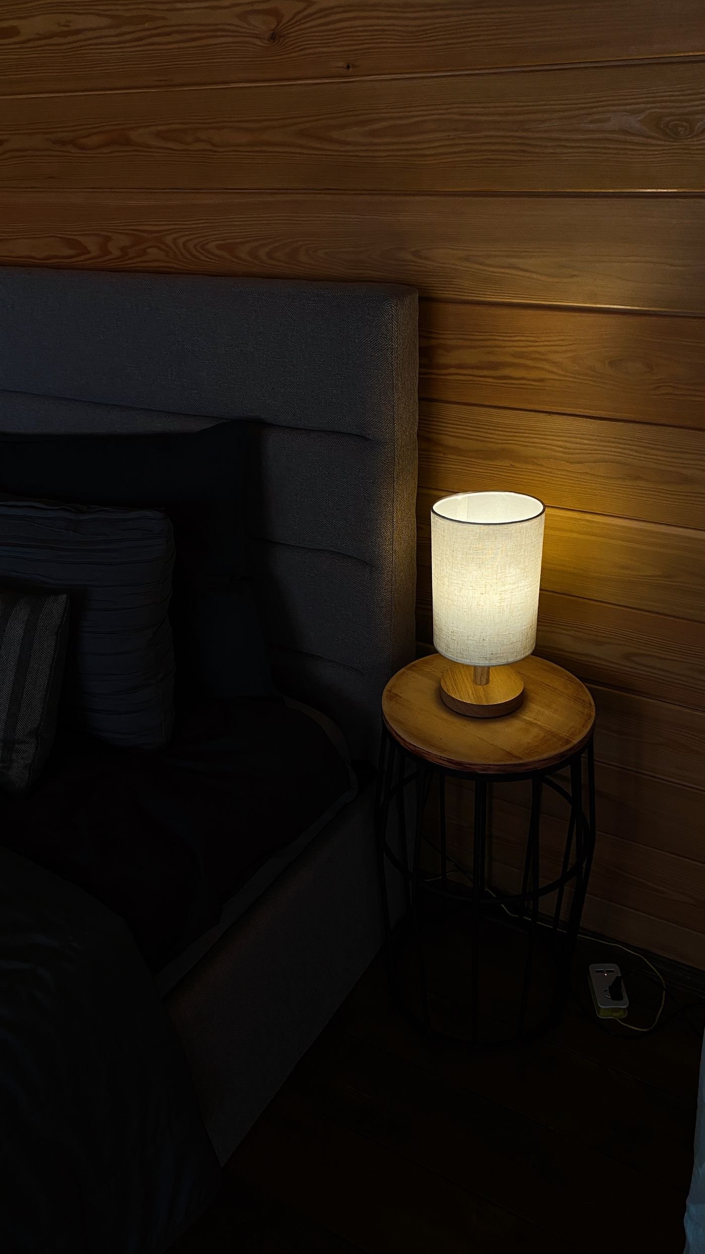 A small warm-lit lamp on a wooden bedside table beside a made bed in an otherwise dark room